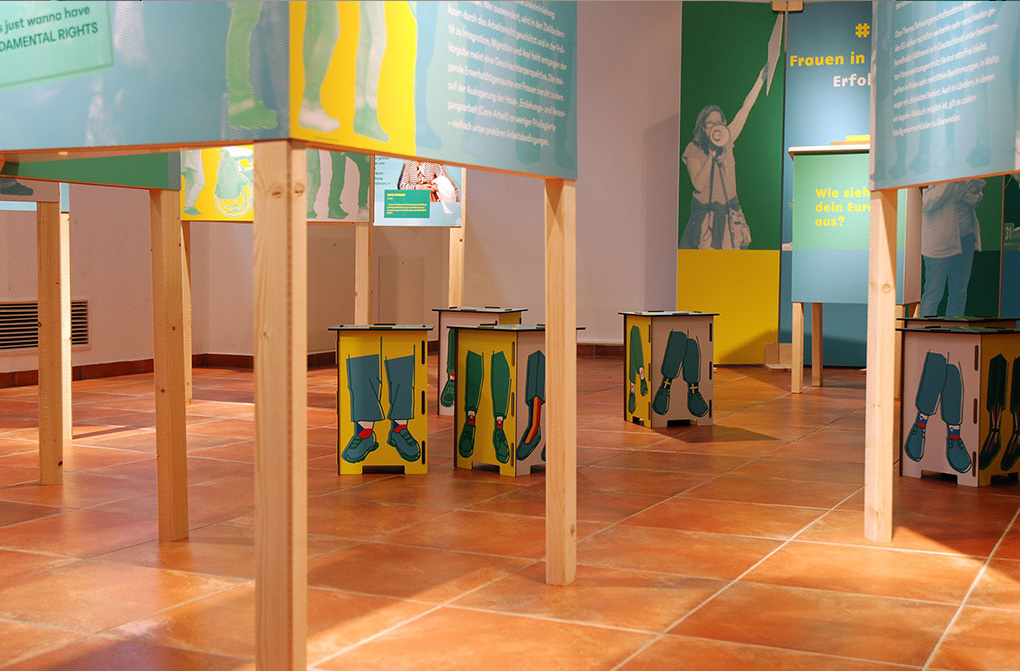 Blick in die Wanderausstellung, mit Fokus auf die Hocker, die Grafiken zu den Mitgliedstaaten der EU zieren / View of the touring exhibition, focussing on the stools adorned with graphics of the EU member states